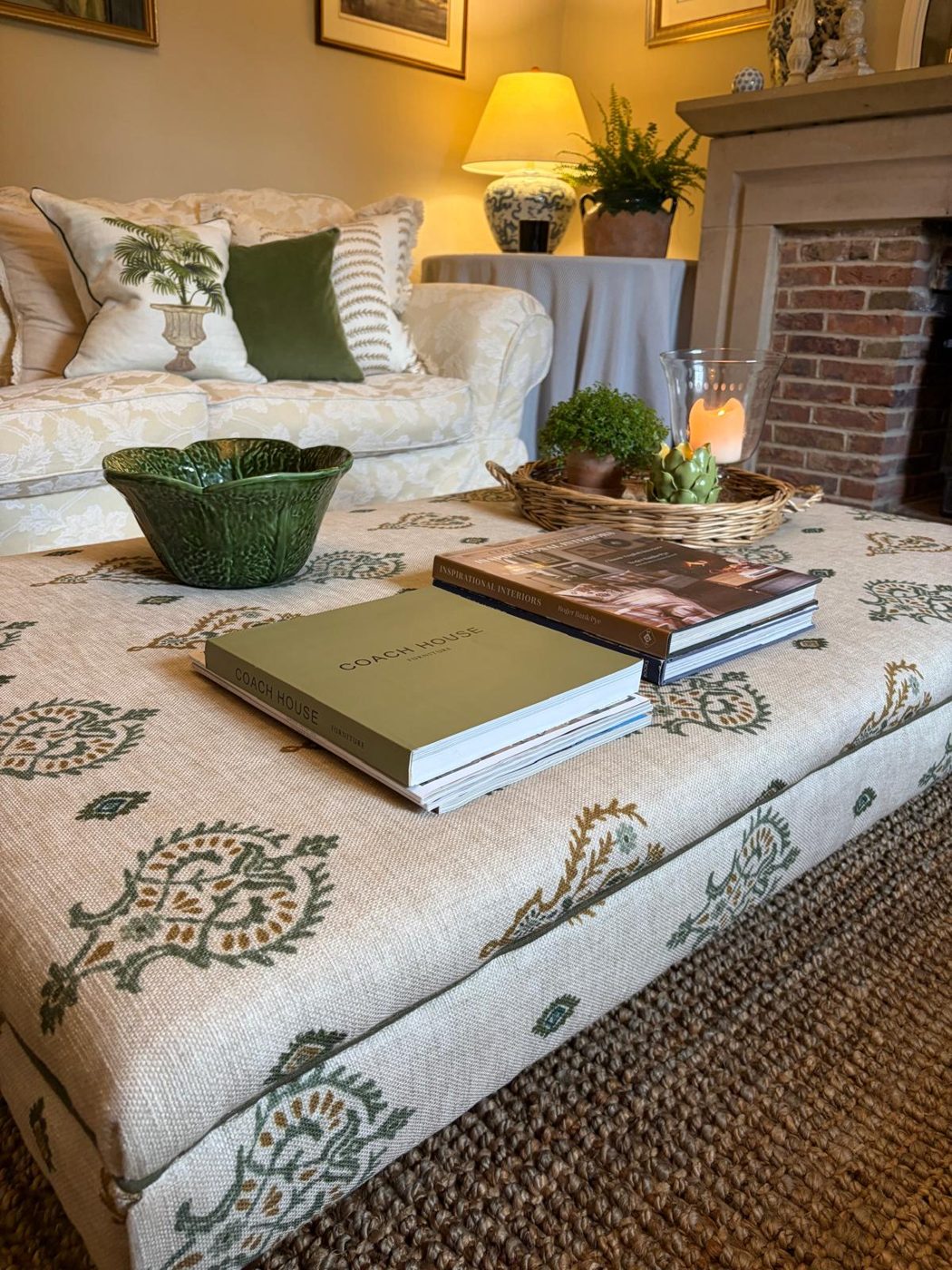 A patterned ottoman in front of a cream sofa with green and palm-print cushions, a green bowl, stacked books and a glowing lamp