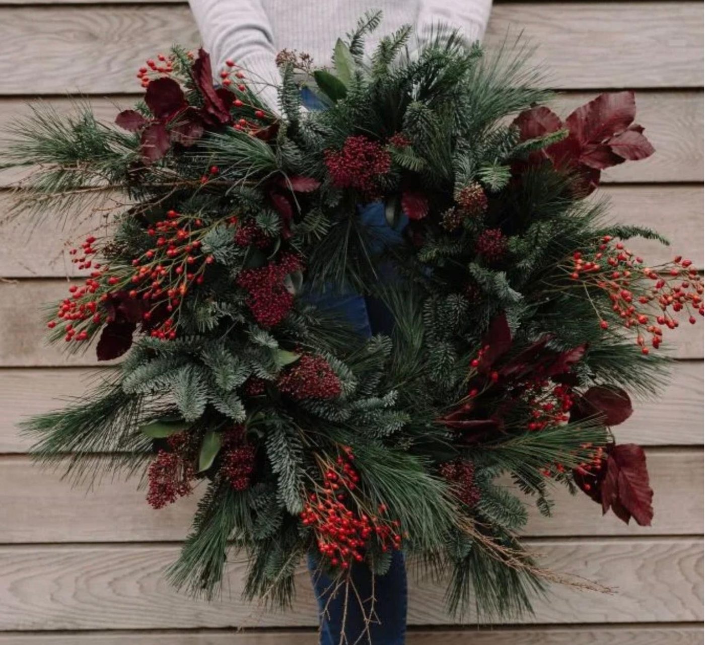 A full natural wreath of evergreen, red rosehips, burgundy leaves and berries held against a wooden wall