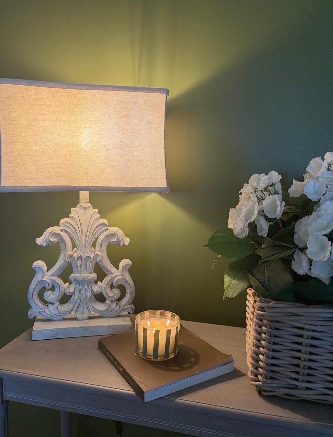 A carved white table lamp glowing against a deep green wall, with a striped candle on a book and white hydrangeas in a wicker basket